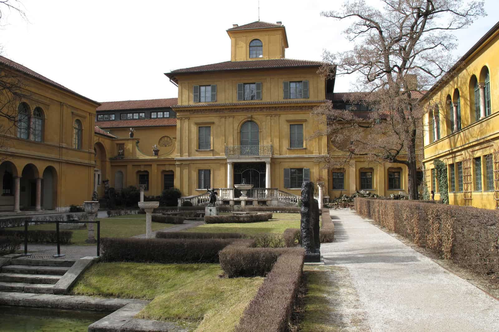 Das historische gelbe Lenbachhaus steht hinter einer steinernen Gartenmauer in München.