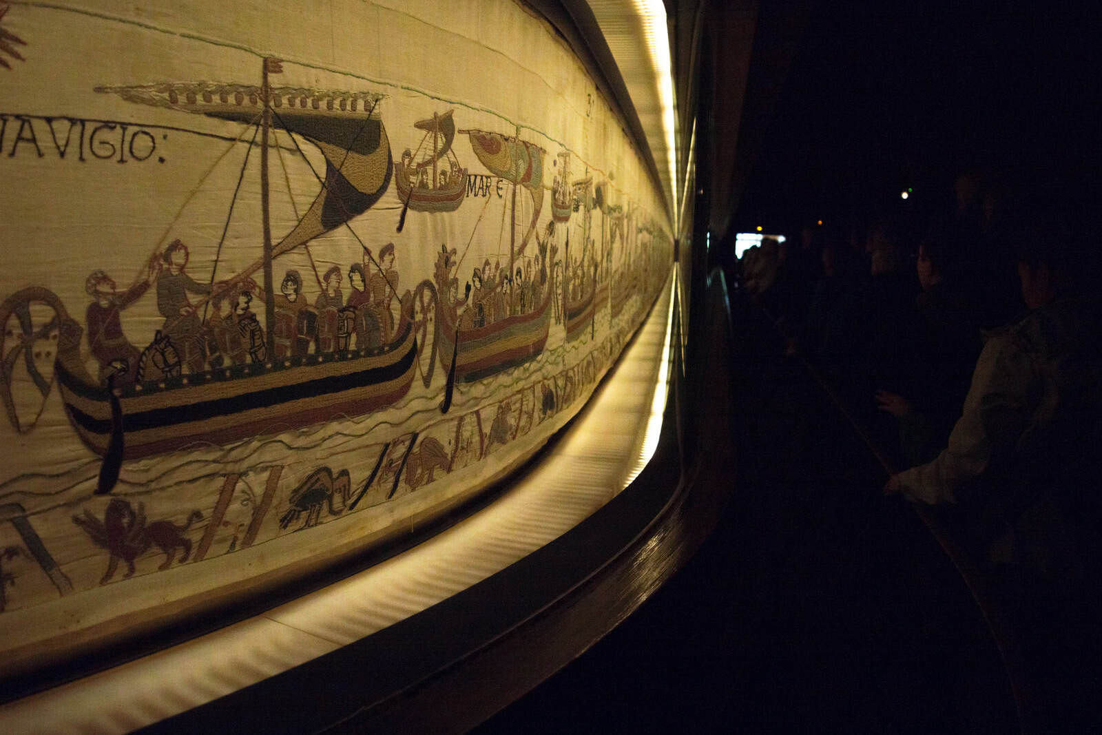 Der Teppich von Bayeux liegt als langer beleuchteter Bildstreifen in einer abgedunkelten Museumsgalerie hinter Glas.